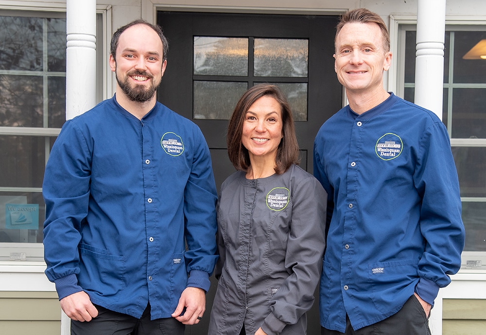 Three dental professionals in branded uniforms stand together outside a dental office building.