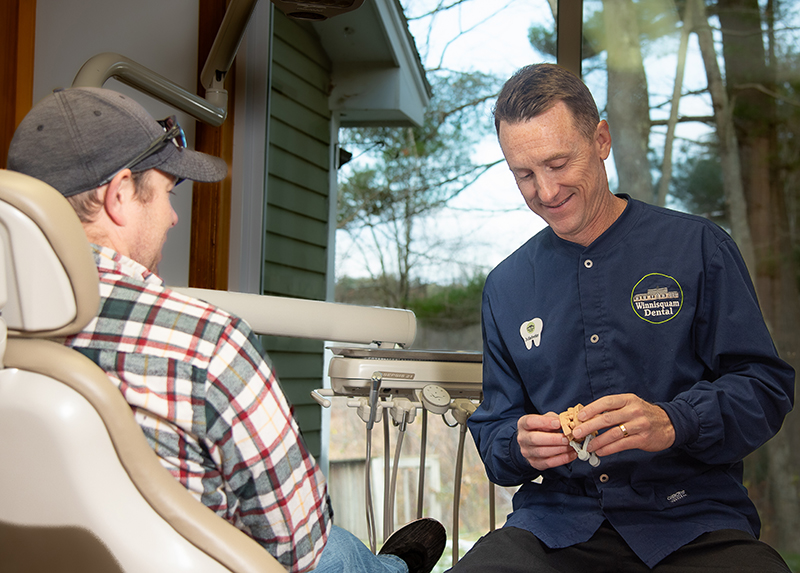 Two men on a porch discussing dental care; one wears plaid shirt, other wears navy dental jacket.