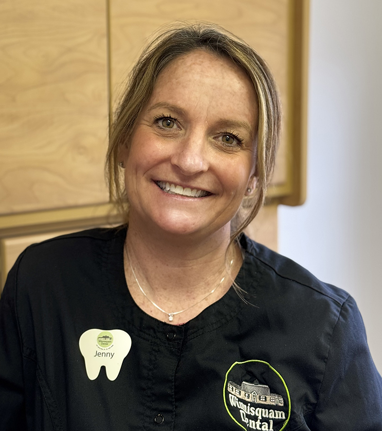 Dental professional smiling at camera wearing black scrubs with tooth pin in clinical office setting.