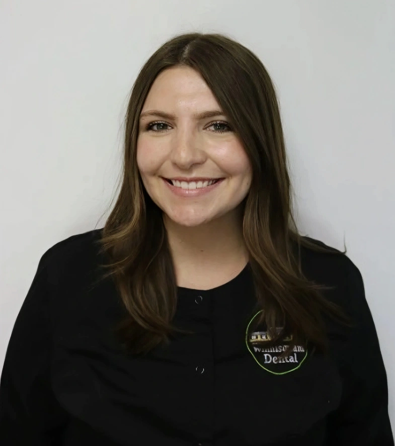 A headshot of a smiling woman with shoulder-length, straight brown hair parted down the middle. She is looking directly forward, wearing a black scrub top featuring a circular green and white logo on the left chest that includes the word "Winnisquam Dental". The background is a solid, plain white.