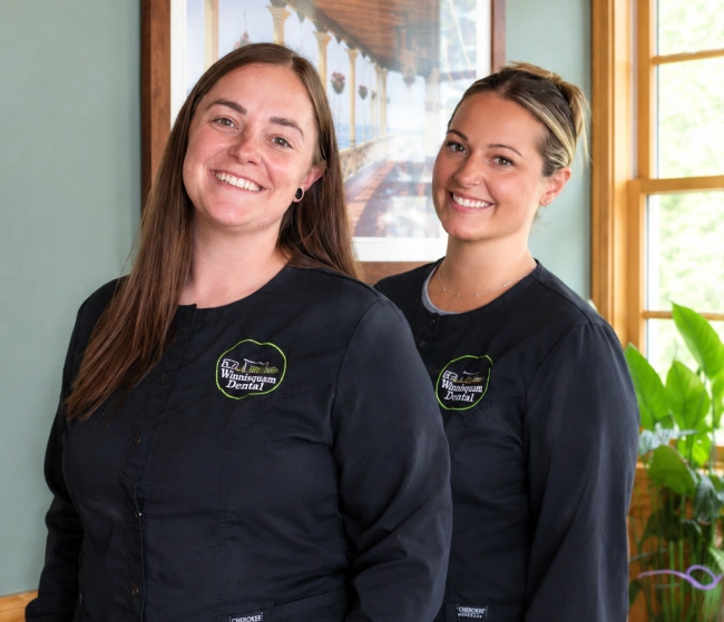 Two smiling Winnisquam Dental staff members in matching black scrub jackets, standing indoors near a window