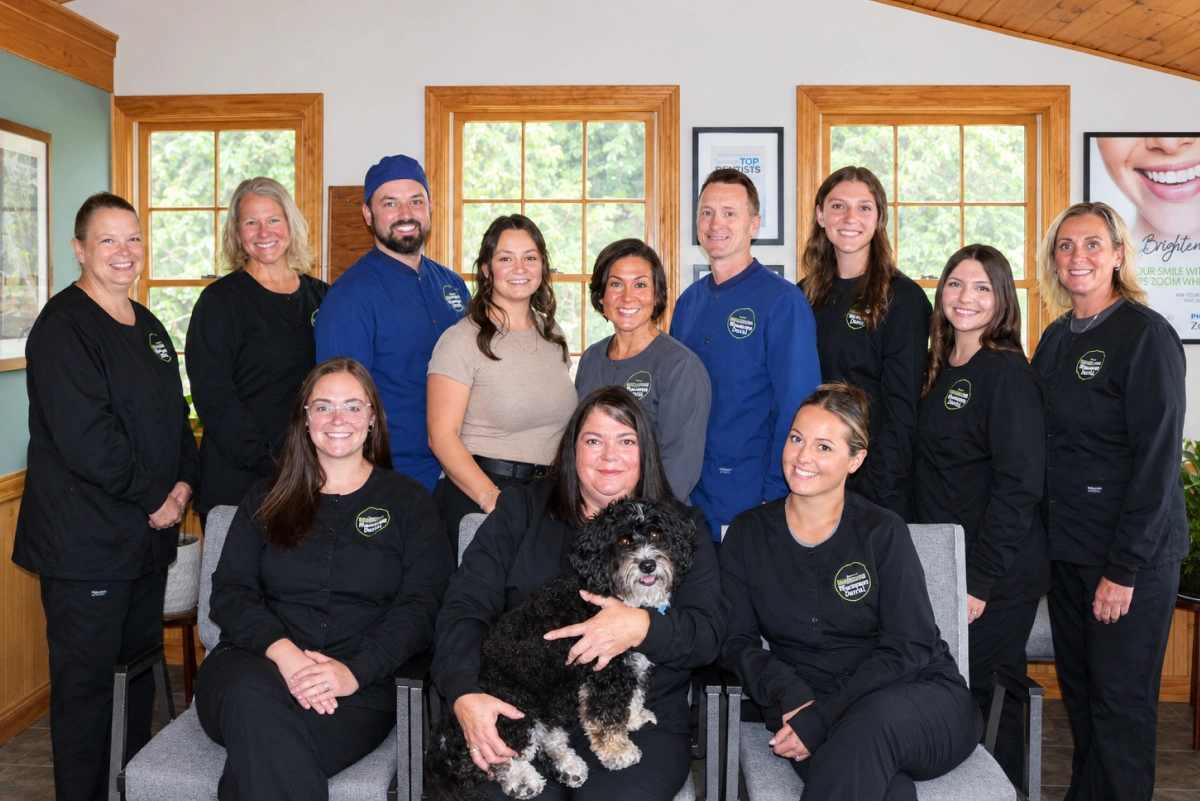 Group photo of the Winnisquam Dental team indoors, most in matching black scrub uniforms, with a dog seated in front